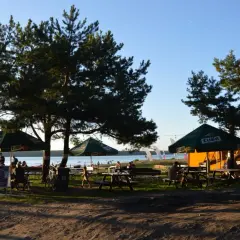 Relaks na plaży Karolinów. Ludzie siedzą pod parasolami, rowery czekają na plaży, a w tle jezioro i namioty.