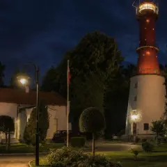 Nocna panorama: charakterystyczna latarnia morska w Rozewiu, biały budynek z czerwonym dachem, ceglany komin i oświetlona zieleń.