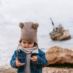 Małe dziecko w czapce z pomponami przygląda się swoim dłoniom na tle wraku statku. To świetne miejsce na odkrywanie przygód, czyli cypr atrakcje dla dzieci.