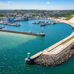 Port w Władysławowie z licznymi łodziami i falochronem, na którym widać spacerujących ludzi. W oddali plaża i zabudowania.