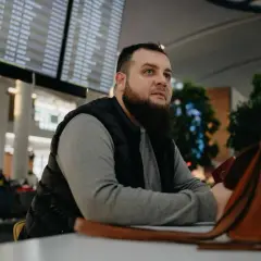 Mężczyzna z brodą siedzi w poczekalni lotniska, patrząc na tablicę odlotów. Czyżby czekał na lot, który jest zagrożony przez overbooking?