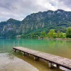 Idealne miejsce na wakacje w **Austrii latem z dziećmi**. Drewniane molo prowadzi do krystalicznie czystej wody, a w tle wznoszą się zielone góry.