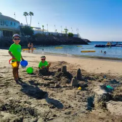 Radosne **teneryfa z dziećmi** na plaży. Dwóch chłopców buduje zamki z piasku, a w tle widać wodne atrakcje.