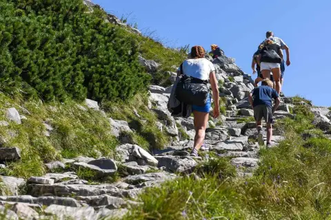 Rodzina wspina się kamienistym szlakiem. Dziecko idzie z przodu, a za nim dorośli. To ich wejście na Giewont.