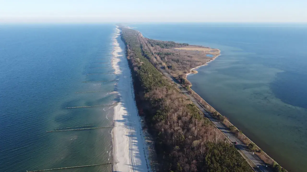 Wąski pas lądu, czyli **półwysep helski**, otoczony przez morze i zatokę. Na plaży widać falochrony, a w głębi las i drogę.