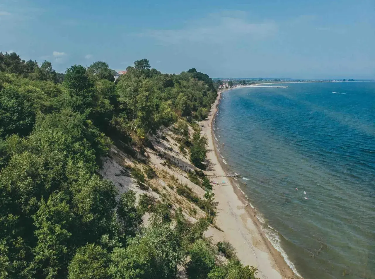 Mechelinki klif porośnięty drzewami, z piaszczystą plażą i turkusowym morzem. W oddali widać zabudowania i falochron.
