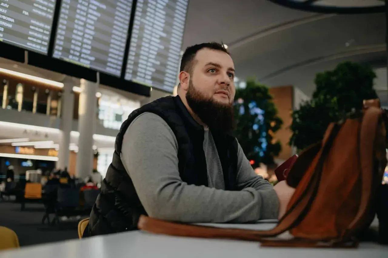 Mężczyzna z brodą siedzi w poczekalni lotniska, patrząc na tablicę odlotów. Czyżby czekał na lot, który jest zagrożony przez overbooking?