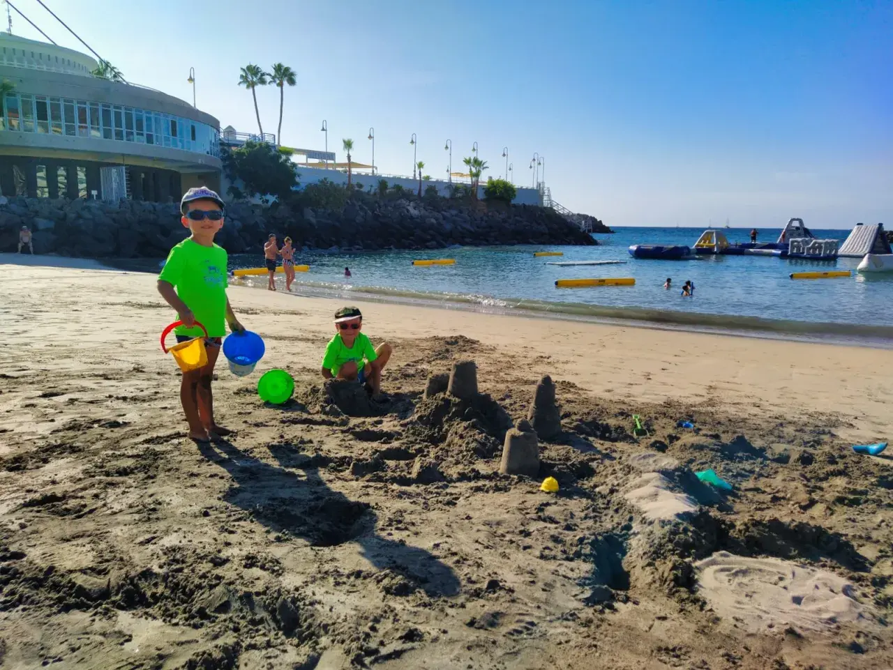 Radosne **teneryfa z dziećmi** na plaży. Dwóch chłopców buduje zamki z piasku, a w tle widać wodne atrakcje.