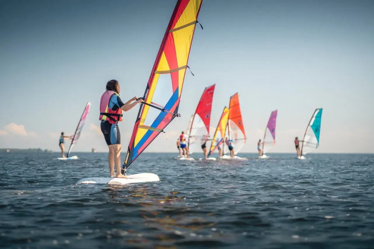 Ludzie na deskach z żaglami, uczący się, co to windsurfing. W tle kolejne kolorowe żagle na wodzie.