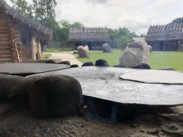 Ognisko pod płaskim kamieniem, na którym piecze się podpłomyk. W tle drewniane chaty i zielony dziedziniec osady średniowiecznej Sławutowo.